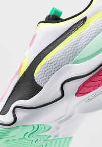 Weißer Sportschuh mit einem strukturierten Mesh-Obermaterial, schwarzen Akzenten, lebhaften gelben, rosa und grünen Details sowie einer flexiblen Gummisohle.