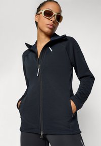 Dunkelblauem Reißverschlussjacke aus strukturiertem Stoff; verfügt über lange Ärmel, seitliche Taschen und einen hohen Kragen. Getragen mit hellen Sonnenbrillen.