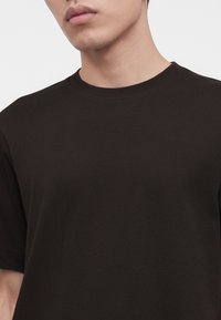 Jeune personne portant un t-shirt noir uni à col rond, visible des épaules jusqu'au milieu de la poitrine, avec une expression neutre.