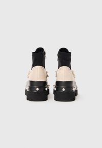 Chunky black and beige ankle boots with platform soles, pearl embellishments around heels, black zippered stretchy upper, shown from the back.