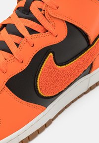 Close-up of an orange and black sneaker with textured orange swoosh, orange laces, and white sole with brown tread.