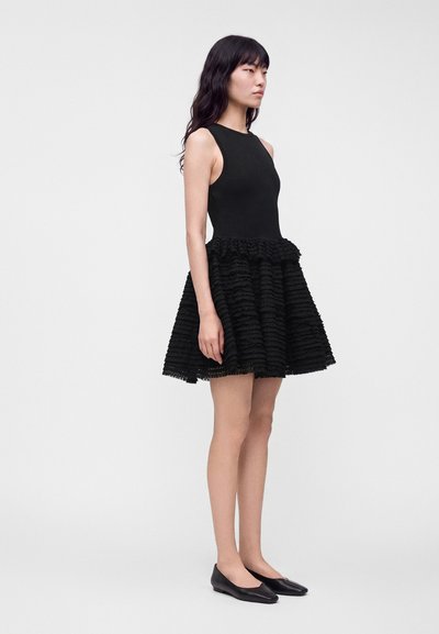 Femme debout dans une robe noire sans manches avec une jupe texturée et à volants, et des chaussures plates noires sur un fond clair uni.