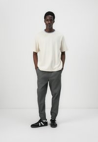 Beige t-skjorte med korte ermer og en blå og lilla ribbestrikket halsringning, kombinert med grå innsnevrede bukser og svarte joggesko med hvite detaljer.