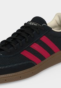 Μαύρο παπούτσι Adidas με κόκκινες ρίγες, μαύρα κορδόνια, υφασμάτινο επάνω μέρος και καφέ λαστιχένια σόλα σε λευκό φόντο.