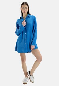 Camicia blu grande e fresca in tessuto texturizzato, con colletto e maniche lunghe, abbinata a sneakers bianche.