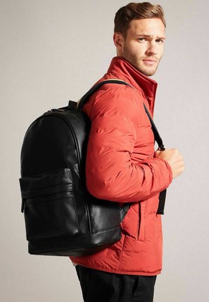 Zaino in pelle nera di forma arrotondata, con tasca frontale e cinghie regolabili, indossato sopra una giacca imbottita rossa.