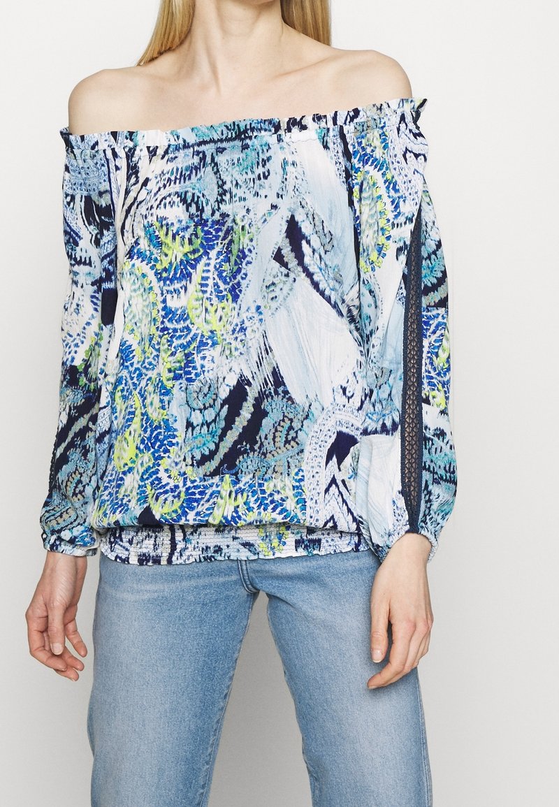 Femme portant un chemisier à épaules dénudées avec un motif abstrait bleu, vert et noir et un jean bleu clair sur fond uni.