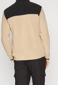 Chaqueta de forro polar beige con un panel superior negro, que presenta mangas largas y un cuello alto. Textura suave y acentos en contraste.