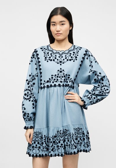 Νεαρή γυναίκα με ανοιχτό μπλε φόρεμα που έχει μαύρη ανθισμένη κεντητική και κυματιστά τελειώματα, στέκεται με το ένα χέρι στη μέση της μπροστά σε απλό φόντο.