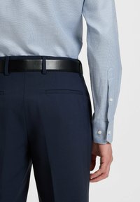 Lichtblauw gemêleerd overhemd met een knoopmanchet, gecombineerd met donkere marineblauwe broek, vastgehouden door een zwarte leren riem. Twee achterzakken zijn zichtbaar.