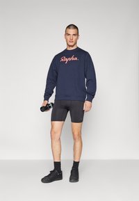 Sudadera azul marino con el logo "Rapha", pantalones cortos de compresión negros y zapatillas negras. Modelo sostiene una botella de agua, de pie frente a un fondo gris.