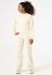 Suéter de punto crema con diseño de trenzas texturizadas, mangas en forma de globo y dobladillo acanalado, combinado con pantalones de campana crema claros y zapatillas.