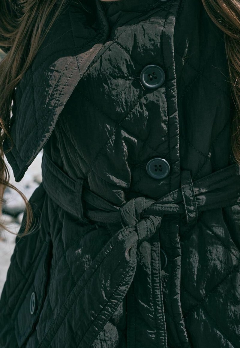 Gros plan d'un manteau matelassé foncé avec des boutons et une ceinture nouée autour de la taille, montrant les détails du tissu texturé.