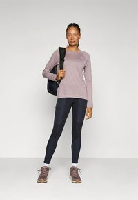 Top de mangas compridas lilás claro, leggings pretas com aplicações em patchwork, sapatilhas desportivas castanhas, segurando uma mala preta. Tecido liso, corte desportivo.