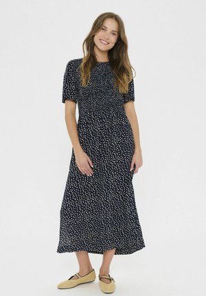 Marineblå, kortærmet maxikjole med en smocket bodice og mønster af hvide polka dots. Skørtet flowet ned til anklerne og er parret med beige flade sko.