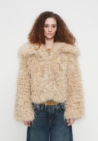 Μπεζ μπουφάν από αφράτη γούνινη συνθετική γούνα με φαρδιά γιακά, υπερμεγέθη μανίκια και κουτί σχήμα. Φορεμένο πάνω από μια μουσταρδί μπλούζα και σε συνδυασμό με μπλε τζιν.