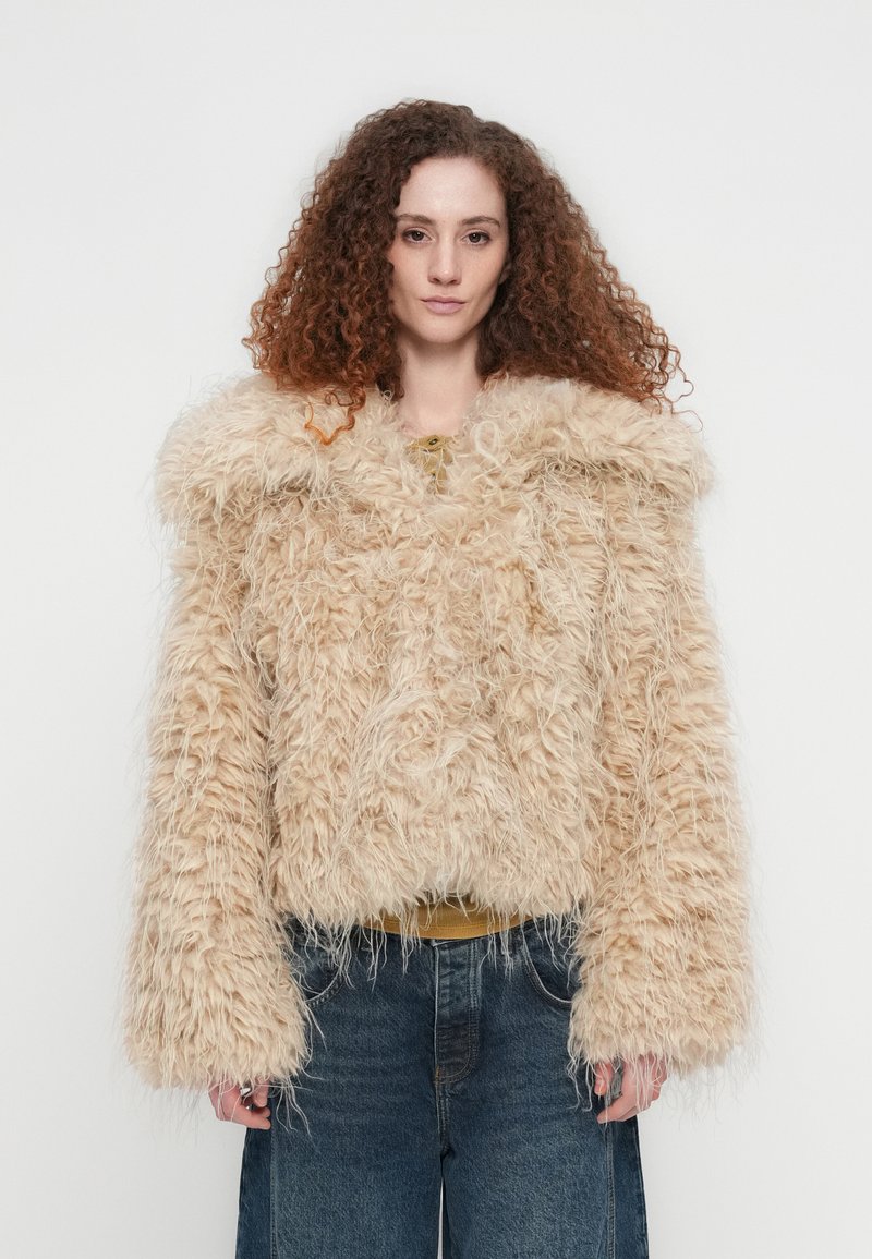 Μπεζ μπουφάν από αφράτη γούνινη συνθετική γούνα με φαρδιά γιακά, υπερμεγέθη μανίκια και κουτί σχήμα. Φορεμένο πάνω από μια μουσταρδί μπλούζα και σε συνδυασμό με μπλε τζιν.