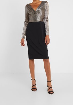 Woman wearing a knee-length black skirt and a long-sleeve metallic silver wrap top with black strappy high heels.