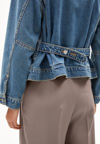 Achteraanzicht van een persoon in een blauwe denimjack met een verstelbare knopenriem en taupe broek. Hand ontspannen aan de zijkant.
