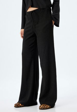Black wide-leg pants with a textured fabric; elastic waistband with drawstring. Paired with brown shoes featuring a leopard print design.