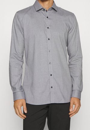 Mężczyzna ubrany w szarą kraciastą koszulę zapinaną na guziki z długim rękawem oraz czarne spodnie, stojący na jednolitym białym tle.