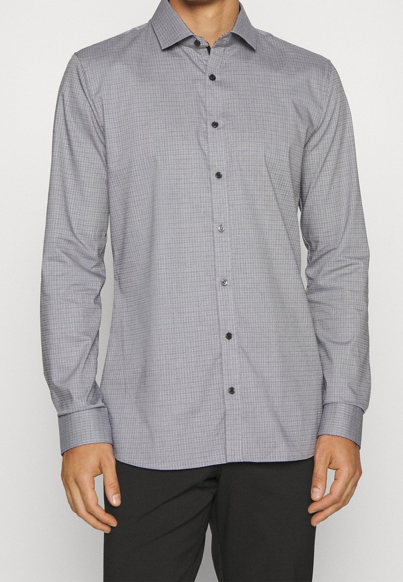Homme portant une chemise à carreaux grise à manches longues boutonnée avec un pantalon noir, debout sur un fond blanc uni.