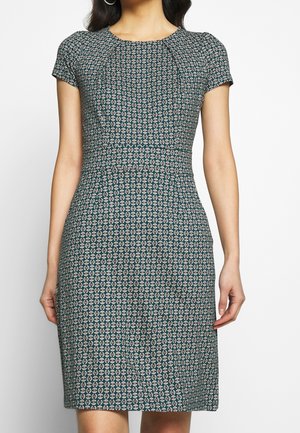 Woman wearing a fitted short-sleeve dress with a geometric circular pattern in dark green and beige tones.