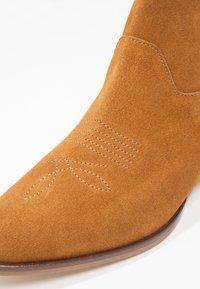 Botte de cowboy en daim beige avec une couture décorative sur l'embout et une semelle en bois, montrée en gros plan.