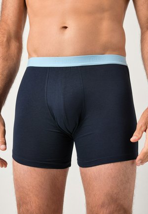 Torso masculin portant un boxer bleu foncé avec une ceinture bleu clair, debout devant un fond neutre.