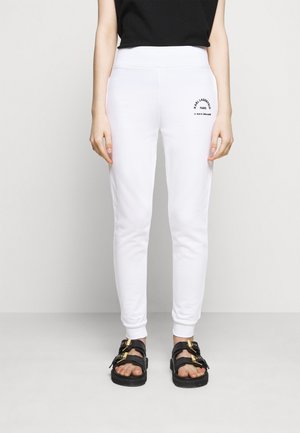 Personne portant un pantalon de jogging blanc fuselé avec un logo noir sur la cuisse et des sandales noires avec des boucles dorées, debout devant un fond uni.