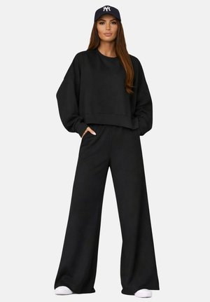 Femme portant un sweat-shirt noir oversized et un pantalon large, avec des baskets blanches et une casquette de baseball bleu marine, debout les mains dans les poches.