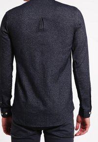 Donkerblauw lange mouwen shirt van gestructureerde stof. Heeft een rugplooitje en knoopsluitingen bij de manchetten. De zoom heeft een afgeronde vorm.