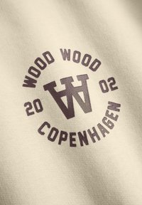Crème katoen stof met een circulair logo met "WOOD WOOD" en "COPENHAGEN" in donkerbruine tekst, opgericht in 2002.