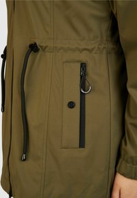 Olivengrüne Jacke mit strukturiertem Finish, ausgestattet mit einer schwarzen Reißverschlusstasche, Knopfdetails und verstellbaren Kordeln am Saum und an den Ärmelbündchen.