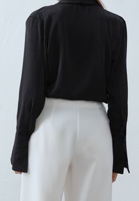 Maglietta nera a maniche lunghe con texture liscia, colletto e polsini con bottone, abbinata a pantaloni bianchi drappeggiati a vita alta.