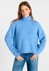 Vrouw in een lichtblauwe, pluizige coltrui en blauwe spijkerbroek, staand met de handen op de heupen tegen een effen witte achtergrond.
