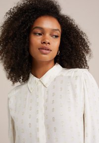 Blusa bianca a maniche lunghe con motivo a pois dorati, colletto strutturato e maniche leggermente a sbuffo. Il tessuto testurizzato aggiunge interesse visivo.