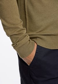 Olivgrüner Pullover mit langen Ärmeln, gerippten Bündchen und lässigem Schnitt, kombiniert mit dunkelblauen Hosen. Subtile Textur im Stoff sichtbar.