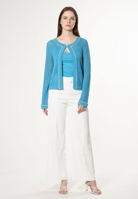 Leichte, blaue Strickjacke mit offenem Gewebe, weißen Akzenten und Rundhalsausschnitt, kombiniert mit einem figurbetonten türkisfarbenen Oberteil und weitem weißen Hosen.