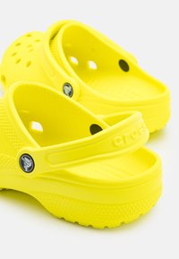 Sabots en plastique jaune avec des trous pour la ventilation, une lanière transversale pour le soutien, une semelle intérieure texturée et un logo circulaire sur le côté.