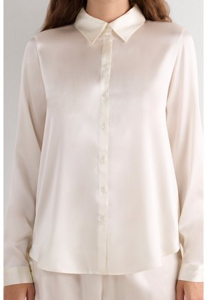 Femme portant une chemise en satin ivoire à manches longues avec boutons et col pointu, debout devant un fond uni clair.