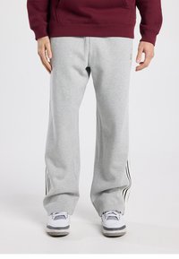 Pantalon de survêtement gris à coupe décontractée, avec des bandes latérales noires et une texture douce. Assorti à des baskets blanches et un sweat à capuche bordeaux.