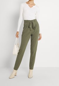 Olivgröna högmidjade byxor med knytmidja, tillsammans med en vit långärmad topp och beige klackkängor.