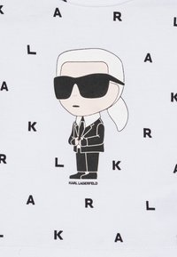 Maglietta bianca con un personaggio dei cartoni animati dai capelli argento e occhiali da sole neri, circondato da testo nero disperso "KARL" e "LAGERFELD".