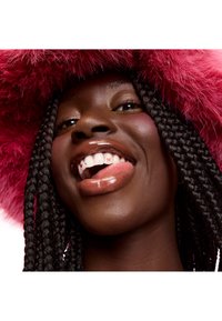 Close-up of a person with braided hair, wearing a fluffy pink hat. They have glossy lips and noticeable dental details with small designs.