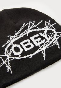Black knit beanie featuring a white graphic with the word "OBEY" encircled by a spiky pattern, creating a distinct contrast and texture.