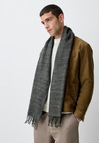 Écharpe à carreaux grise en tissu doux, avec des extrémités effilochées. Portée sur un pull en tricot crème et une veste en daim marron, complétant une palette de couleurs neutres.