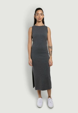 Donna con lunghi capelli scuri che indossa un vestito midi grigio scuro senza maniche e sneakers bianche, in piedi contro uno sfondo chiaro e uniforme.