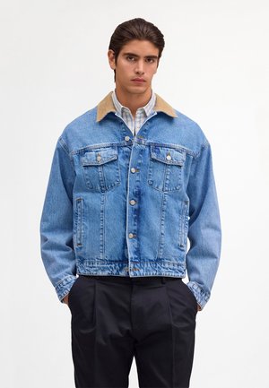 Młody mężczyzna w jasnoniebieskiej kurtce jeansowej z beżowym kołnierzem, nałożonej na koszulę w kratę i czarne spodnie, stoi z rękami w kieszeniach na białym tle.