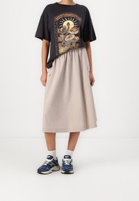 Sort grafisk T-shirt med kunstillustration, beige knælang nederdel og blå sneakers med hvide detaljer. Tekstureret stof og afslappet pasform.